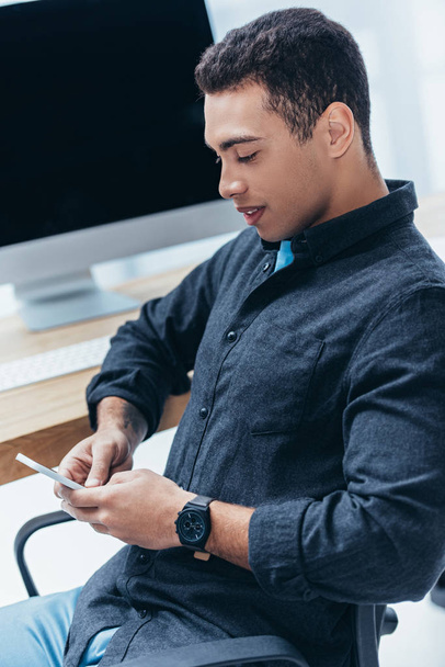 korkea kulma näkymä hymyilevä nuori liikemies istuu ja käyttää älypuhelimen toimistossa
  - Valokuva, kuva