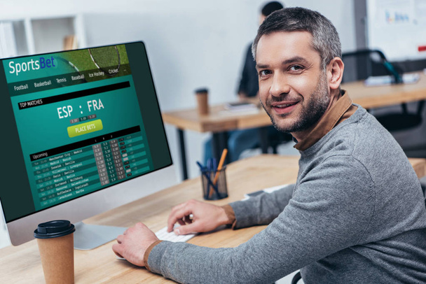 handsome bearded businessman working with desktop computer with sports bet website and smiling at camera in office - Φωτογραφία, εικόνα