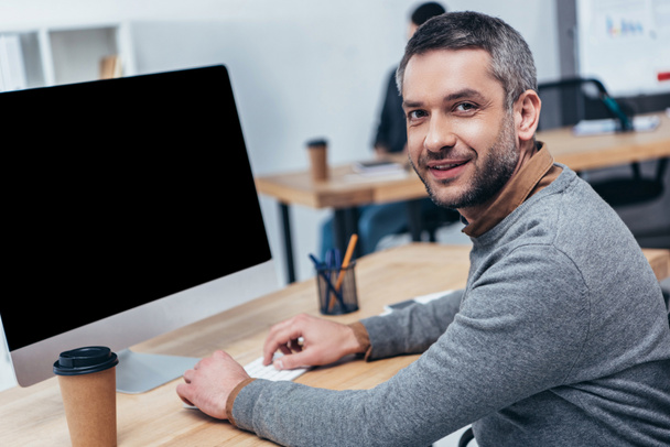 handsome bearded businessman working with desktop computer and smiling at camera in office - Foto, immagini