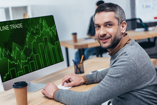 handsome bearded businessman working with desktop computer with online trade graphs and smiling at camera in office - 写真・画像