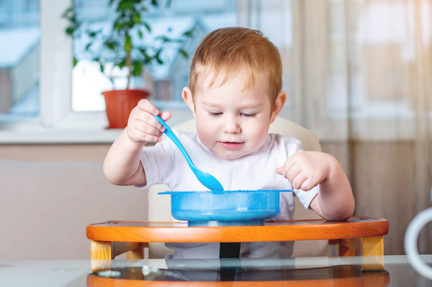 小さな男の子は台所で子供たちのテーブルで自らスプーンですくって食べることを学ぶします。赤ちゃんの健康食品の概念 - 写真・画像