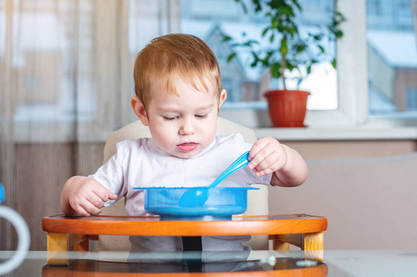 小さな男の子は台所で子供たちのテーブルで自らスプーンですくって食べることを学ぶします。赤ちゃんの健康食品の概念 - 写真・画像