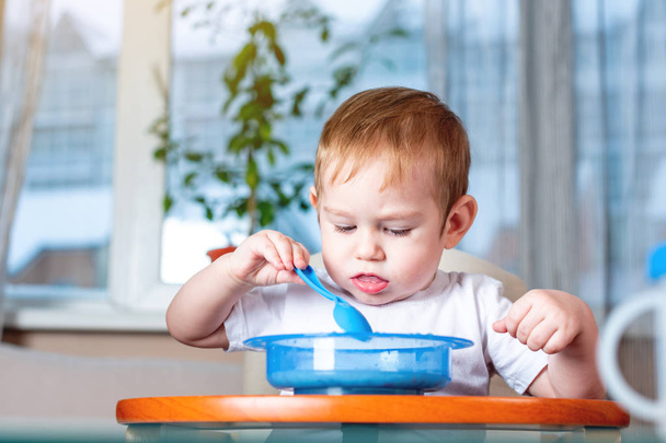 Pequeño niño aprendiendo a comer con una cuchara en la mesa de los niños en la cocina. El concepto de comida saludable para bebés
 - Foto, Imagen