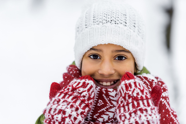 vista da vicino del bambino afroamericano in maglia cappello, guanti e sciarpa sorridente e guardando la fotocamera in inverno
 - Foto, immagini