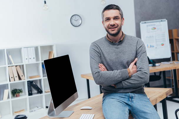 handsome bearded businessman sitting with crossed arms on desk and smiling at camera in office - Zdjęcie, obraz