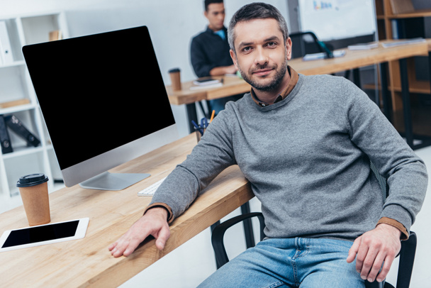 handsome man looking at camera while sitting at table with desktop computer and digital tablet in office - Photo, Image