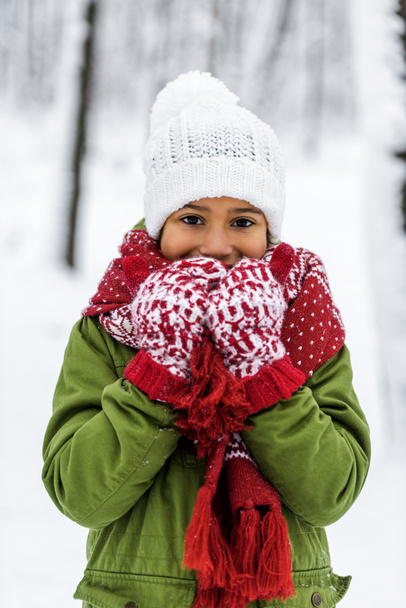 african american child in knitted hat, mittens and scarf smiling and looking at camera in winter park - Photo, Image