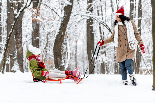 attractive african american woman pulling daughter on sledge in winter park - Foto, afbeelding