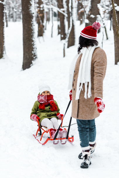 moeder kijken naar Afro-Amerikaanse dochter in slee in winter park - Foto, afbeelding