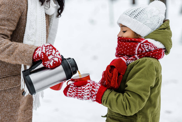 cropped view of mother pouring tea from thermos in red cup holding by african american daughter in winter - Photo, Image