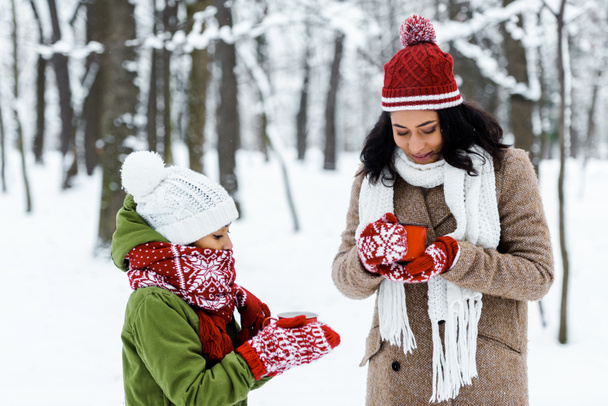 attractive african american mother and cute daughter holding cups of tea in snowy forest - Photo, Image