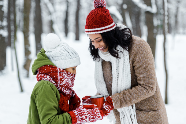 attractive african american mother and cute daughter holding cups of tea in snowy forest - Photo, Image