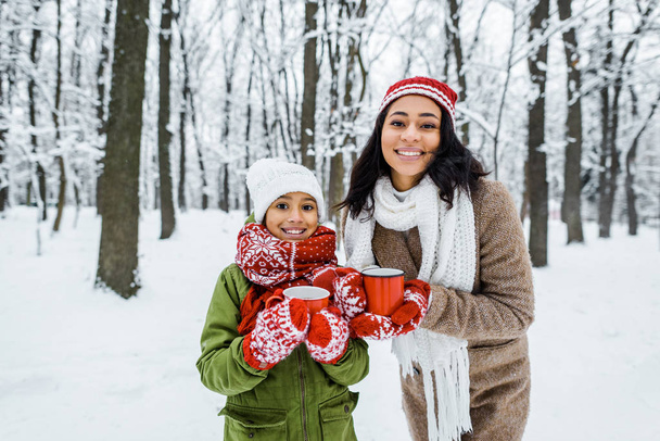 attractive african american mother and cute daughter holding cups of tea and smiling while looking at camera in snowy forest - Valokuva, kuva