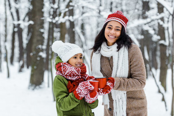 attractive african american mother and cute daughter holding red cups of tea and staying near trees in snowy forest - Photo, Image