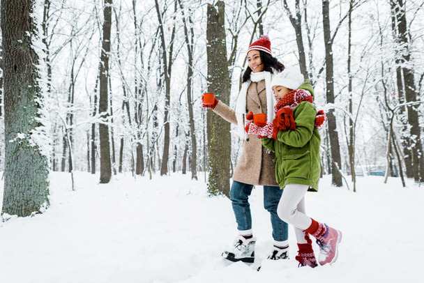 attractive african american mother and cute daughter walking and holding red cups between trees and looking at snow in snowy forest - Photo, Image