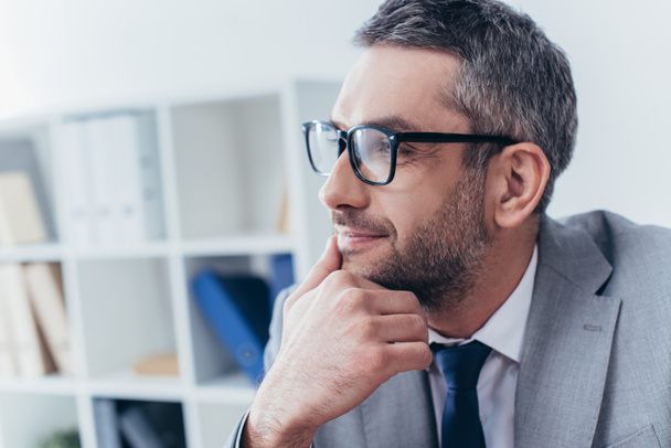 handsome bearded businessman in eyeglasses holding hand on chin and looking away in office  - Photo, Image