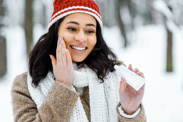 attraente donna afroamericana toccando guancia e applicando crema protettiva nella foresta invernale
 - Foto, immagini