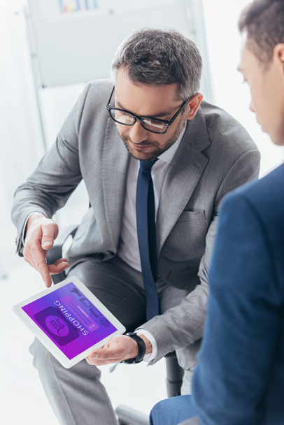 обрезанный снимок бизнесмена в очках, показывающий цифровой планшет с приложением для покупок в Интернете коллеге по работе
 - Фото, изображение
