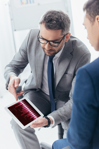 cropped shot of businessman in eyeglasses showing digital tablet with trading graphs to male colleague in office - Foto, imagen