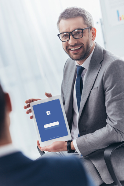 smiling businessman in eyeglasses holding digital tablet with facebook app on screen - Foto, Imagen