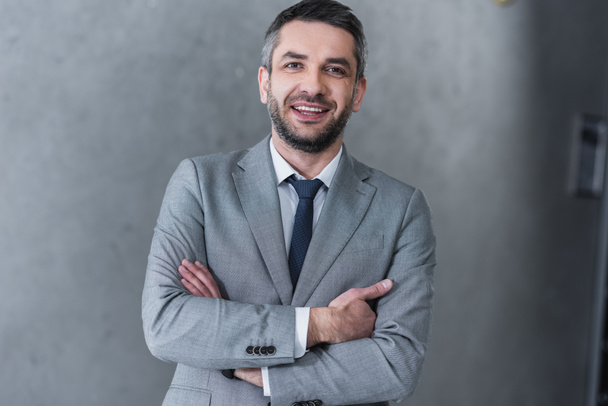 confident handsome businessman standing with crossed arms and smiling at camera - Zdjęcie, obraz