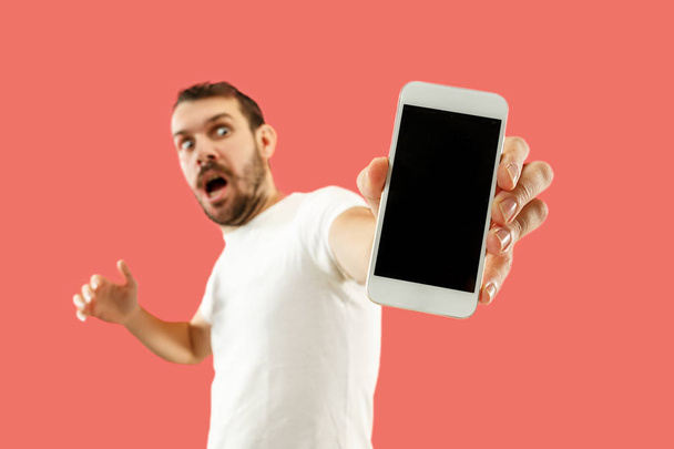 Young handsome man showing smartphone screen isolated on coral background in shock with a surprise face - Foto, Imagen