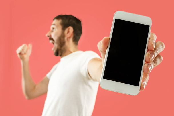 Young handsome man showing smartphone screen isolated on coral background in shock with a surprise face - 写真・画像