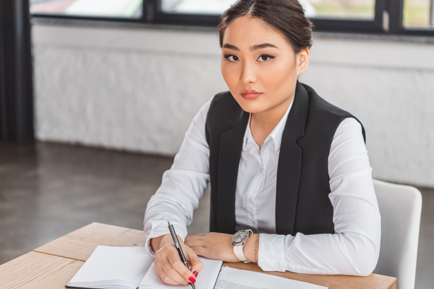serious asian businesswoman writing in notebook and looking at camera in office - Photo, Image