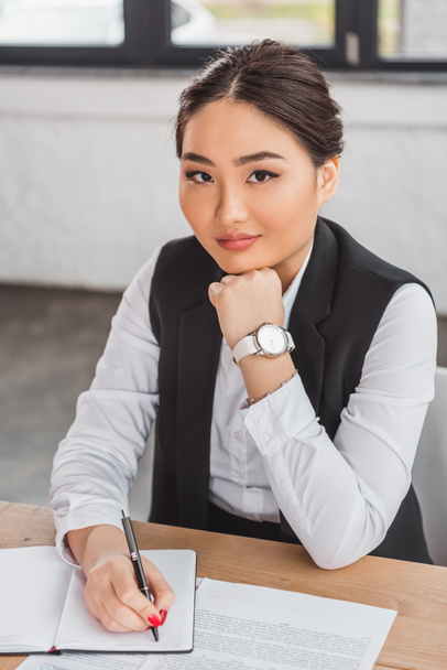 gyönyörű fiatal ázsiai üzletasszony álla és kamera irodában mosolyogva ül - Fotó, kép