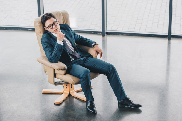 alto angolo vista di fiducioso asiatico uomo d'affari in occhiali seduta con mano sul mento in ufficio
  - Foto, immagini