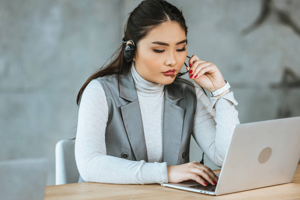 joven asiático mujer de negocios en auriculares usando laptop en lugar de trabajo
 - Foto, Imagen