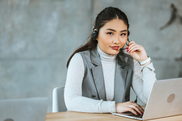 young businesswoman in headset using laptop and looking at camera in office - Foto, immagini