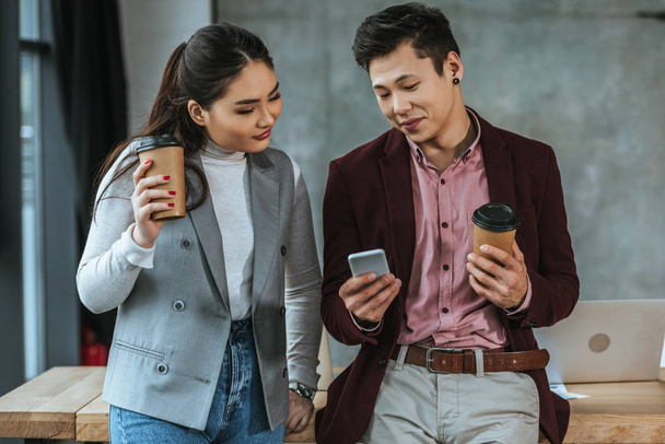 happy young asian business colleagues holding coffee to go and using smartphone in office - 写真・画像