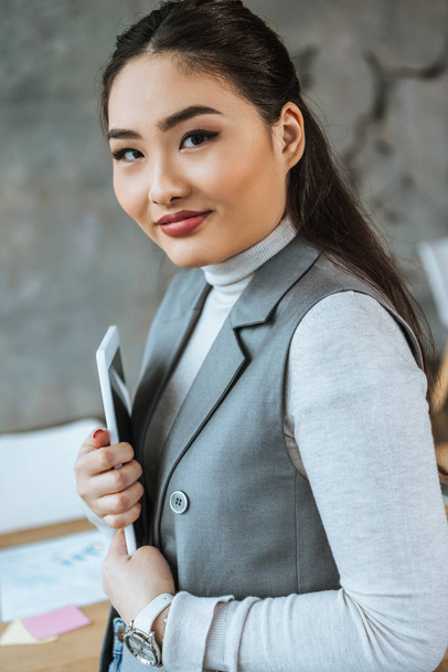 hermosa joven asiático mujer de negocios celebración digital tableta y sonriendo a la cámara
  - Foto, Imagen