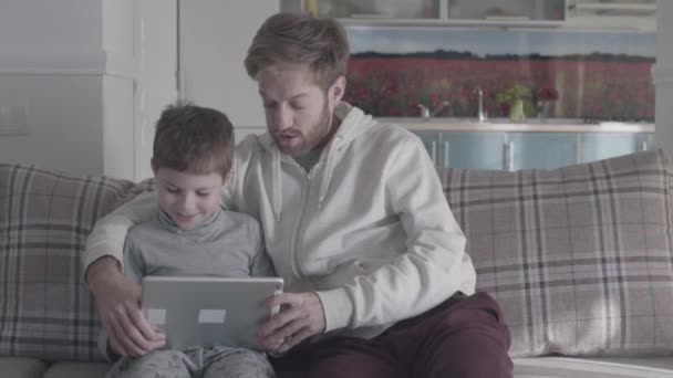 Bearded father and kid using digital tablet sitting on the couch in large living room. Daddy teaches his son. Happy family spend time together. Father-son relationship. - Metraje, vídeo