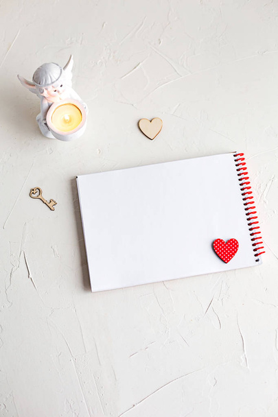 Top view of an open notebook with red pencil and two wooden hearts and a key on white table. Angel candleholder decorations. - Fotoğraf, Görsel
