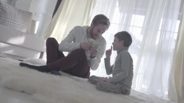 Playful beard father sitting with his son at the carpet and holding money. Dad and kid put a finger to the nose. Joyful daddy counts money. Father-son relationship. - Video