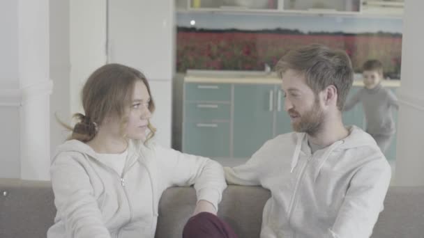 Beautiful couple have conversation sitting on the couch. Little son comes and hugs both parents. Happy family waving hands. Family relationships. - Materiał filmowy, wideo