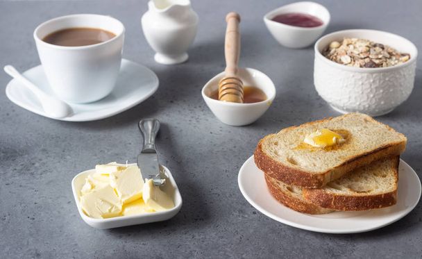 Butter and bread for breakfast with cup of coffee, granola, honey and jam over grey stone background.  - Foto, afbeelding