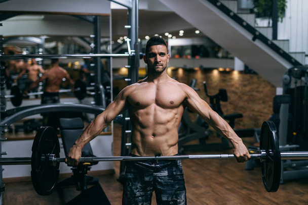 Young strong Caucasian shirtless bodybuilder lifting barbells. Gym interior. - Фото, изображение