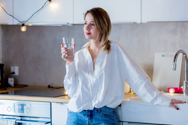 Молодая кавказка в белой рубашке и синих джинсах со стаканом воды останавливается на кухне
. - Фото, изображение