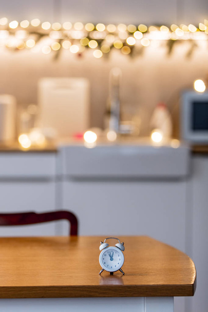 little alarm clock on a table with Christmas lights on background. Kitchen interior - Φωτογραφία, εικόνα