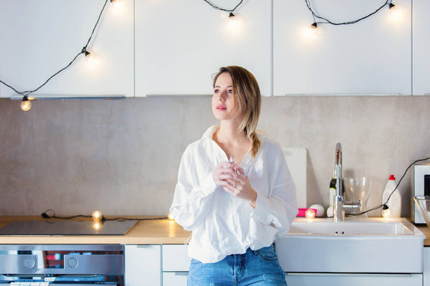 Молода біла жінка в білій сорочці і блакитні джинси зі склянкою води зупиніться на кухні
. - Фото, зображення