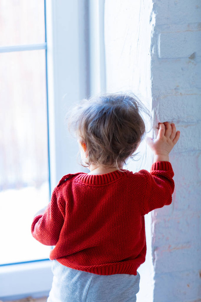 El niño pequeño se queda cerca de la pared y mira a la ventana. Ubicación del hogar
  - Foto, Imagen