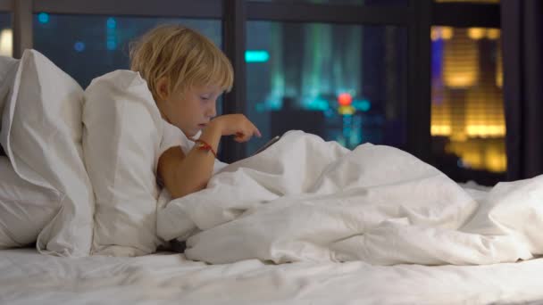 Little blond boy sitting in bed at night and playing digital tablet with silhouettes of city in background. - Filmagem, Vídeo