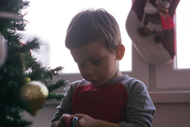 Χαριτωμένο παιδί διακοσμεί το οικογενειακό χριστουγεννιάτικο δέντρο του, ενώ διαδίδει το πνεύμα των Χριστουγέννων στην οικογένειά του. - Φωτογραφία, εικόνα