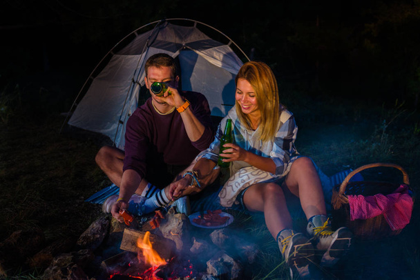 Man and woman baking sausages on the campfire, drinking beer and relaxing by the campfire in the mountain top in the night. Romantic campfire concept. - Photo, Image