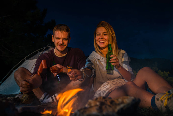 Mann und Frau grillen Würstchen am Lagerfeuer, trinken Bier und entspannen sich abends am Lagerfeuer auf dem Berggipfel. Romantisches Lagerfeuerkonzept. - Foto, Bild