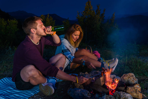 Man and woman rosting sausages on the campfire, drinking beer and relaxing by the campfire in the mountain top in the night. Romantic campfire concept. Motion blur - Photo, Image