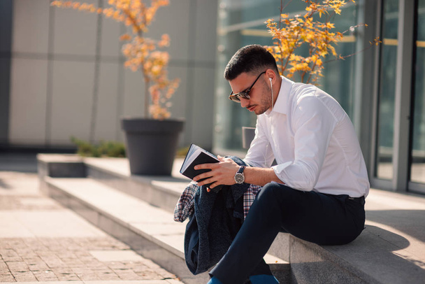 An eloquent businessman is outside, he is enjoying in the beautiful weather while looking at the notebook. Urban businessman on the move. - Фото, изображение